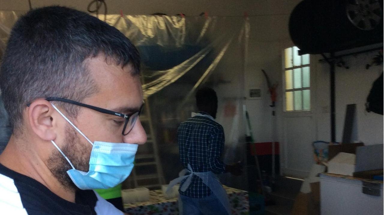 Ein junger Mann mit Brille und Atemschutzmaske steht in der Küche, im Hintergrund ein Mann afrikanischer Herkunft. Ein junger Mann mit Brille und Atemschutzmaske steht in der Küche, im Hintergrund ein Mann afrikanischer Herkunft.