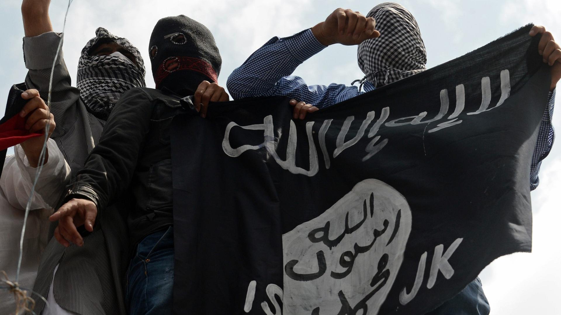 Mehrere Vermummte halten eine schwarze IS-Fahne.