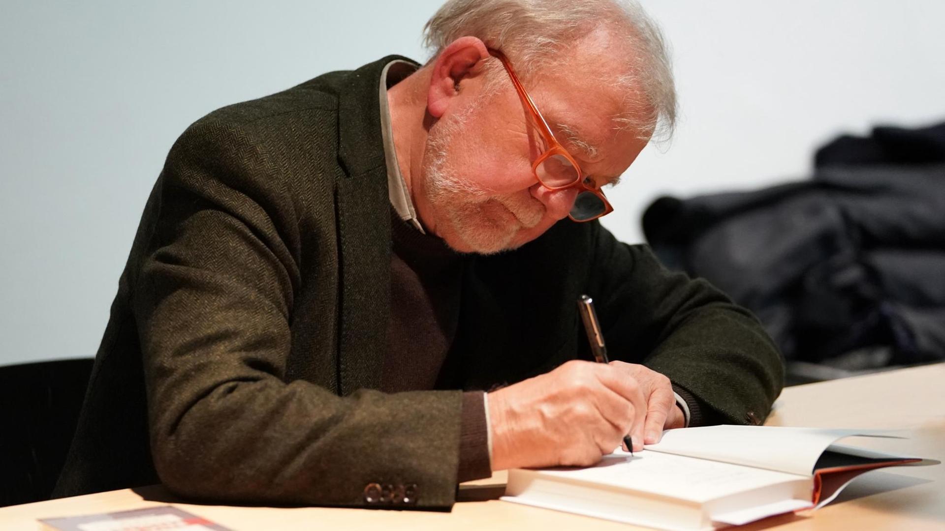 Rüdiger Safranski sitzt bei einer Lesung an einem Tisch und schreibt in ein Buch.