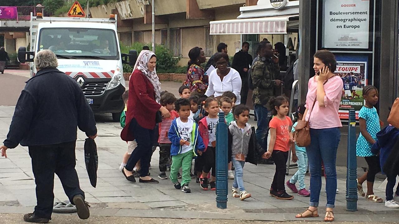 Schulkinder werden von ihren erwachsenen Begleitern durch die Straßen gelotst - eine der Mütter trägt ein Kopftuch Schulkinder werden von ihren erwachsenen Begleitern durch die Straßen gelotst - eine der Mütter trägt ein Kopftuch