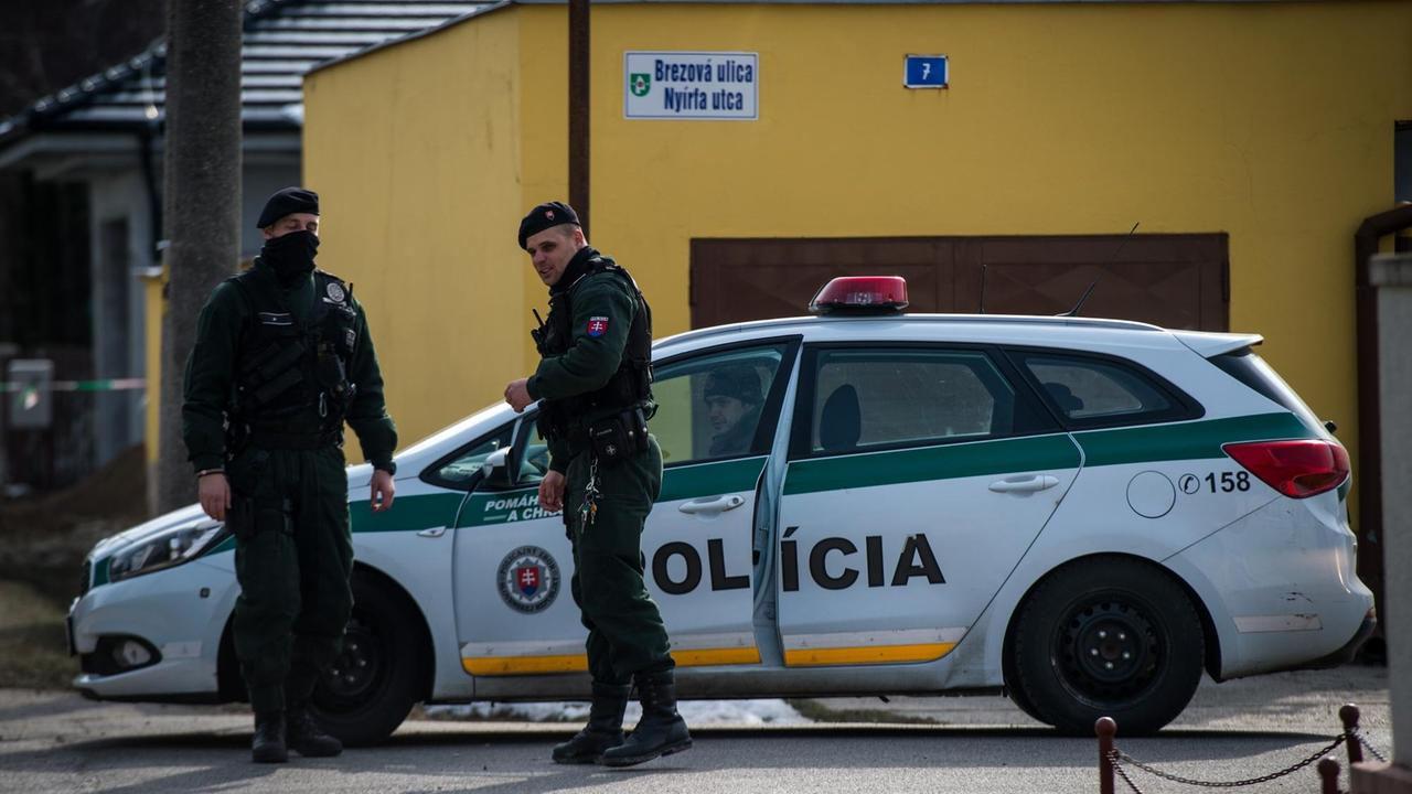 Zwei Polizisten und ein Polizeiauto stehen vor einem gelben Haus. Zwei Polizisten und ein Polizeiauto stehen vor einem gelben Haus.