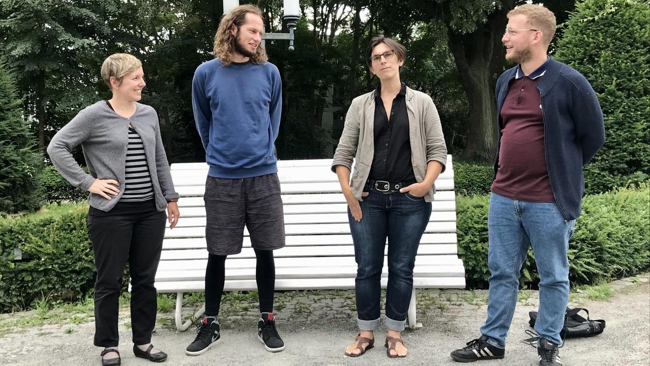 Zwei Männer und zwei Frauen stehen vor einer Bank im Park. Sie schauen Merete Peetz in der Mitte an.