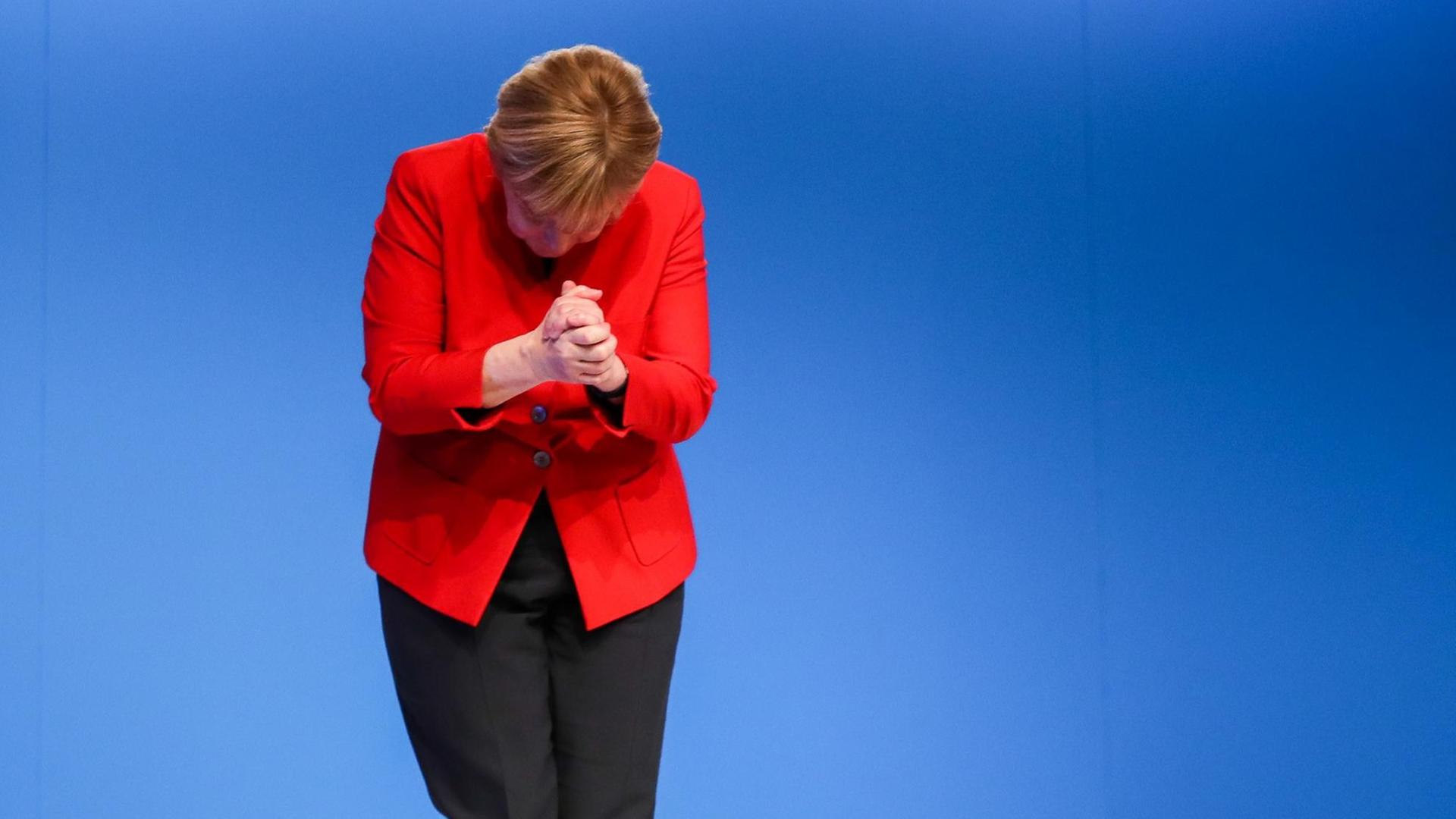 Merkel in roter Jacke vor blauer Wand bei einer Verbeugung Merkel in roter Jacke vor blauer Wand bei einer Verbeugung