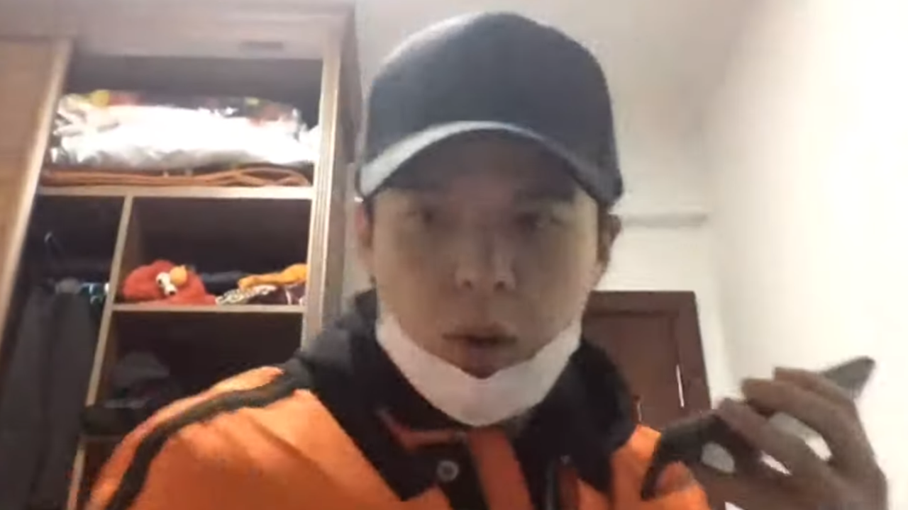 Li Zehua in orangener Jacke, schwarzer Kappe und mit heruntergezogener Atemmaske, in der Hand ein Handy. Er sitzt in einem Raum und spricht in die Kamera.