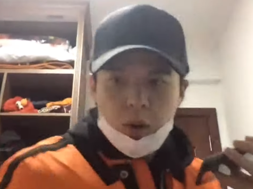 Li Zehua in orangener Jacke, schwarzer Kappe und mit heruntergezogener Atemmaske, in der Hand ein Handy. Er sitzt in einem Raum und spricht in die Kamera.