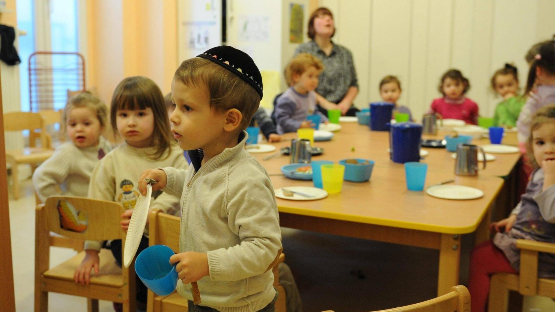 Ein Junge mit einer Kipa trägt einen Teller und einen Becher an einem Tisch vorbei, an dem andere Kinder sitzen. Ein Junge mit einer Kipa trägt einen Teller und einen Becher an einem Tisch vorbei, an dem andere Kinder sitzen.