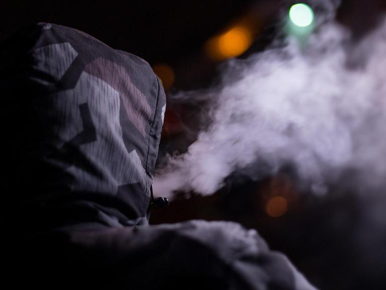 Ein Mann mit hochgezogener Kapuze ist von hinten beim Rauchen zu sehen.