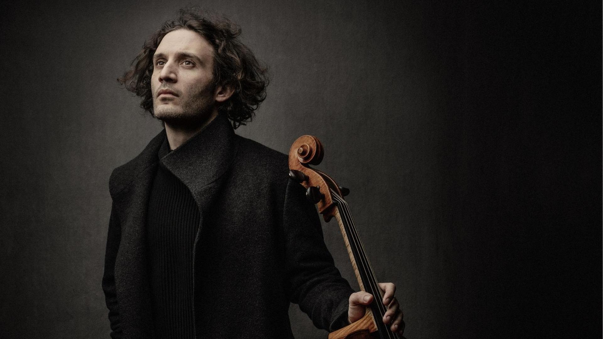 Ein Mann mit mittellangen, dunklen Haaren steht in dunkler Kleidung vor einem dunklen Hintergrund und hält ein Cello in seiner linken Hand.
