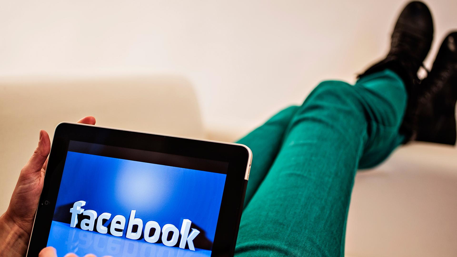 Ein Internetnutzer hat ein Tablet auf dem Schoß, darauf ist eine Facebook-Illustration zu sehen. Seine Beine sind über eine Sofalehne geschwungen. Ein Internetnutzer hat ein Tablet auf dem Schoß, darauf ist eine Facebook-Illustration zu sehen. Seine Beine sind über eine Sofalehne geschwungen.