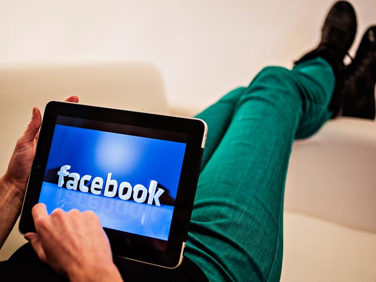Ein Internetnutzer hat ein Tablet auf dem Schoß, darauf ist eine Facebook-Illustration zu sehen. Seine Beine sind über eine Sofalehne geschwungen.