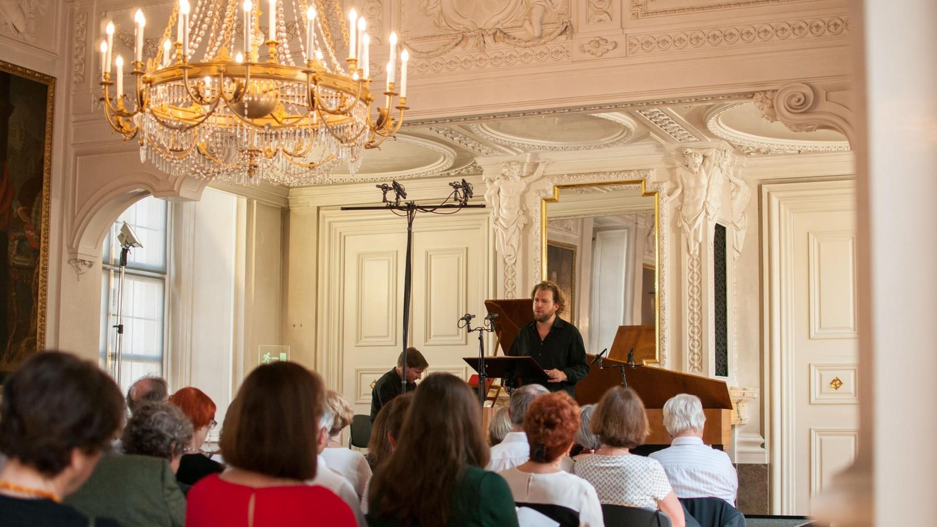 Auf der Bühne steht ein Mann, der singt, und ein Pianist sitzt an einem Klavier. Sie treten in einem herrschaftlichen Saal auf. Auf der Bühne steht ein Mann, der singt, und ein Pianist sitzt an einem Klavier. Sie treten in einem herrschaftlichen Saal auf.