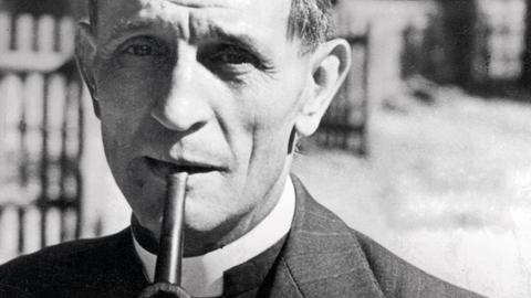 Eine historische Aufnahme von Martin Niemöller, evangelischer Pastor und Theologer, im Anzug und Pfeife im Mund.