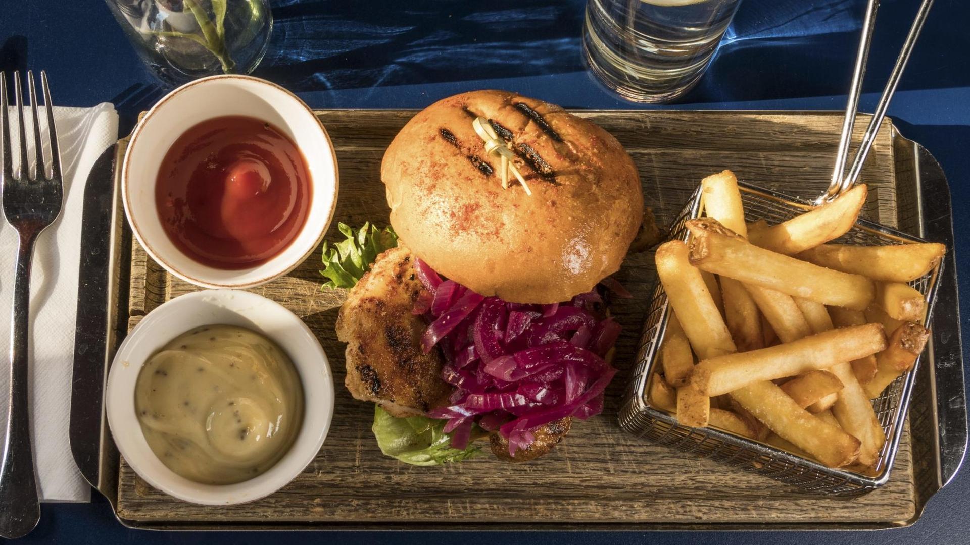 Das Bild zeigt einen Burger mit Hähnchenfleisch und eine Portion Pommes sowie diverse Saucen. Das Bild zeigt einen Burger mit Hähnchenfleisch und eine Portion Pommes sowie diverse Saucen.