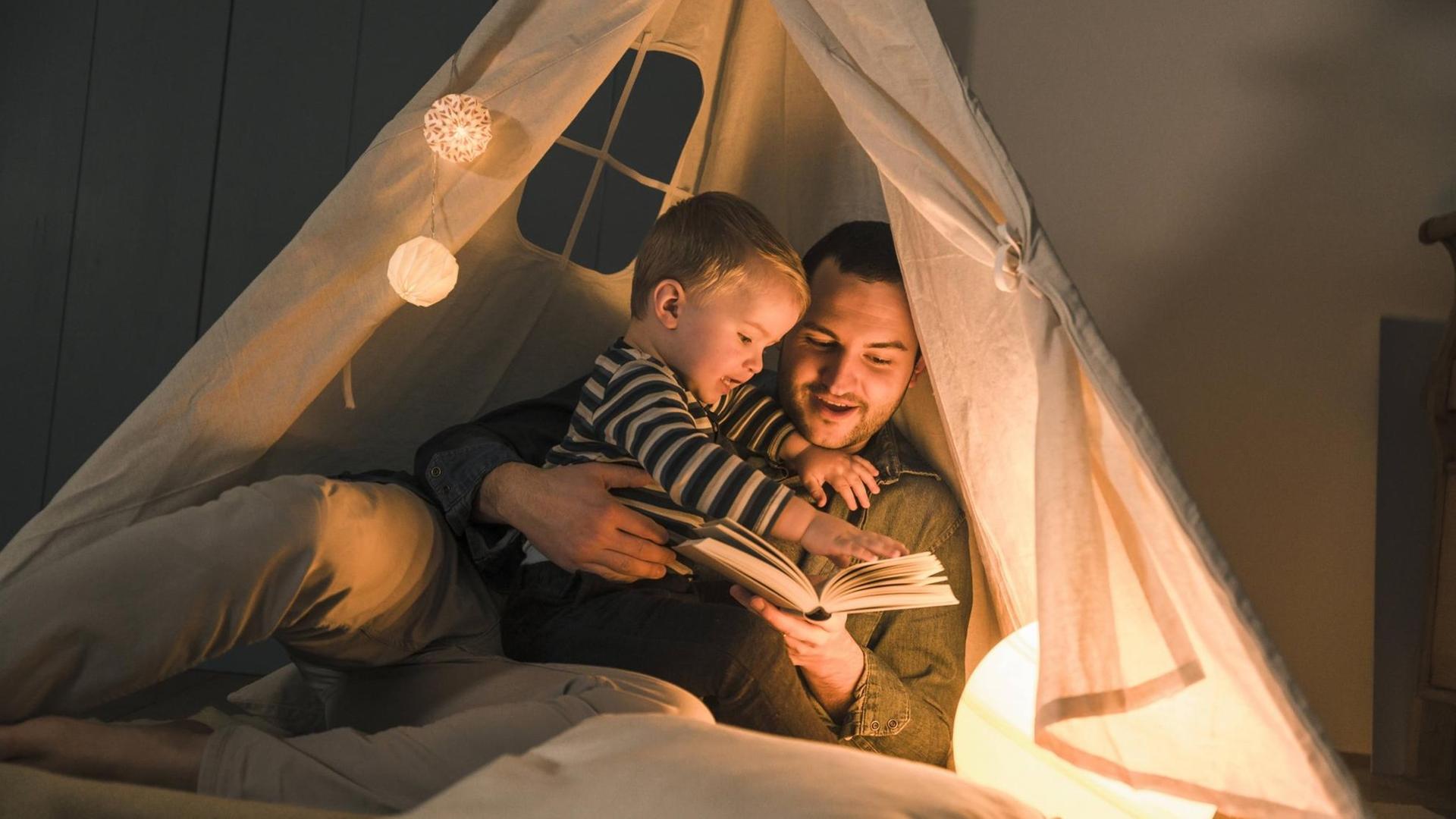 Ein Mann liest seinem Sohn in der Wohnung vor. Beide haben sich in ein Zelt aus weißem Stoff zurückgezogen, das von einer Lampe von innen beleuchtet wird, der Rest der Wohnung ist dunkel.