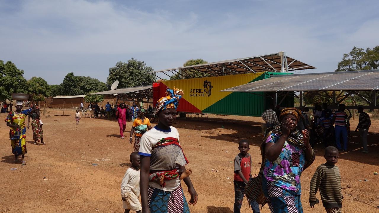 Der Container mit der mobilen Solaranlage steht mitten im Dorf Kai. Auf der Straße gehen Frauen und Kinder vorbei.