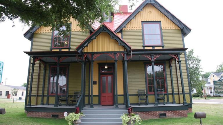Yellow wooden house with black beams.  The roof, windows, front door and some decorations are red.  In front is a typical southern porch.
