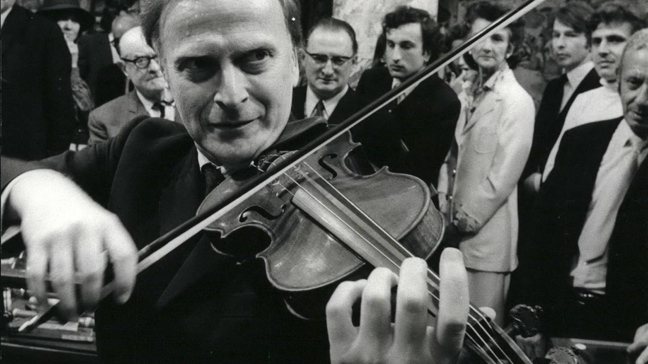 Yehudi Menuhin spielt Geige, während andere im Hintergrund um ihn stehen und zuhören. Yehudi Menuhin spielt Geige, während andere im Hintergrund um ihn stehen und zuhören.
