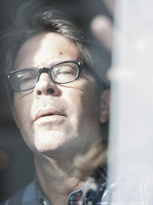 Der US-Autor Jonathan Franzen schaut aus einem Fenster und schiebt dabei eine Gardine beiseite. Er trägt eine Brille.
