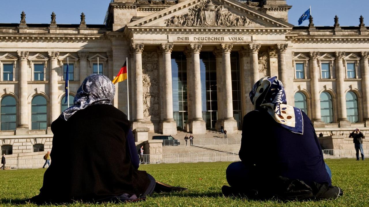 Zwei Frauen mit Kopftuch sitzen auf der Wiese vor dem Reichstagsgebäude...</p>

                        <a href=