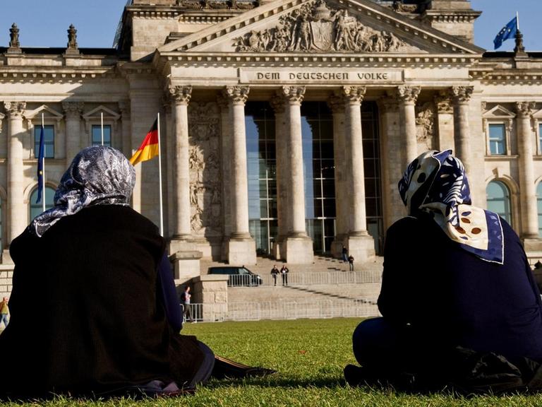 Zwei Frauen mit Kopftuch sitzen auf der Wiese vor dem Reichstagsgebäude in Berlin.