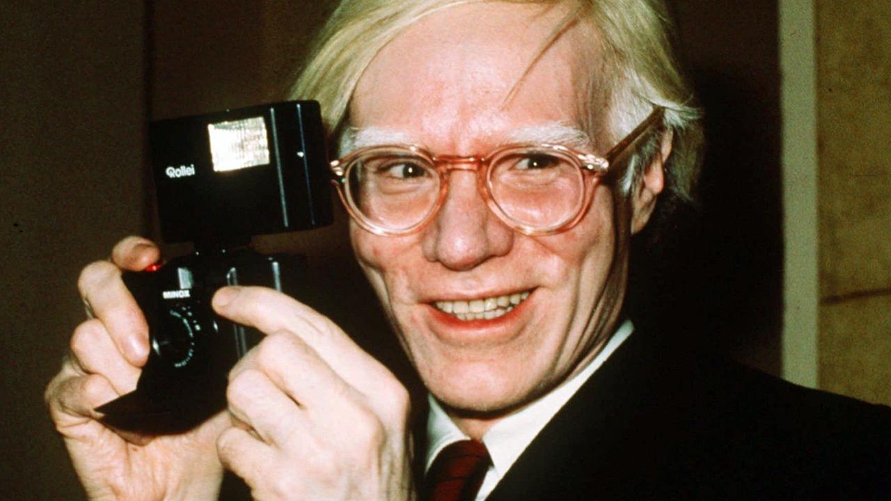 Das Fot zeigt einen lächelden Andy Warhol mit Rollei Kamrea in den Händen.