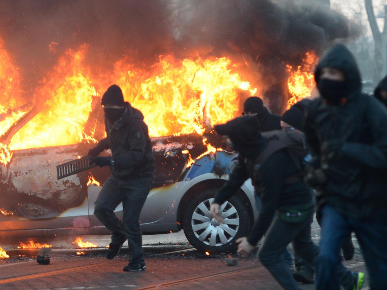 Demonstranten mit schwarzen Kapuzen und Schals, im Hintergrund brennt ein Polizei-Auto Demonstranten mit schwarzen Kapuzen und Schals, im Hintergrund brennt ein Polizei-Auto