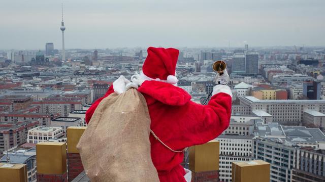 Ein als Weihnachtsmann verkleideter Berufskletterer trägt für eine Werbeaktion auf einem Hochhaus-Dach an Berlins Potsdamer Platz einen Sack voller Geschenke und läutet mit einer Handglocke. Foto: Gregor Fischer/dpa | Verwendung weltweit