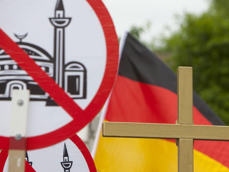 Auf dem Bild sind Schilder mit einer durchgestrichenen Moschee zu sehen, außerdem Deutschlandflaggen und ein Kreuz.