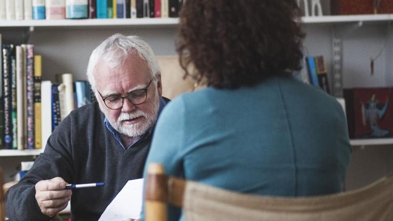 Vor einem gefüllten Bücherregal sitzt ein Therapeut, der im Gespräch mit einer Frau ist. Vor einem gefüllten Bücherregal sitzt ein Therapeut, der im Gespräch mit einer Frau ist.