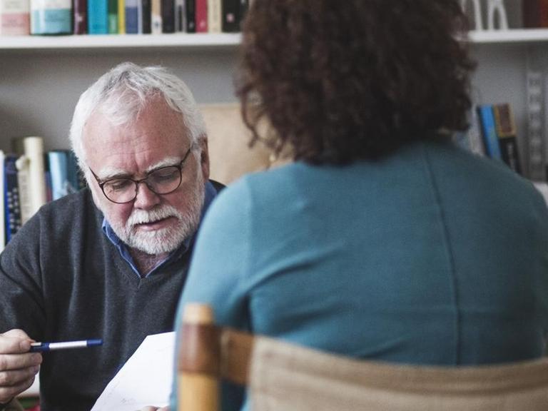 Vor einem gefüllten Bücherregal sitzt ein Therapeut, der im Gespräch mit einer Frau ist.