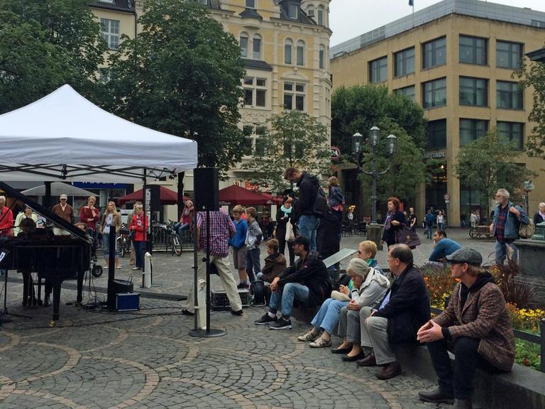 Auf dem Münsterplatz in der Bonner Innenstadt sitzen eine Reihe von Menschen und warten. Ein Flügel ist aufgebaut, überdacht mit einem Zelt gegen den Regen.
