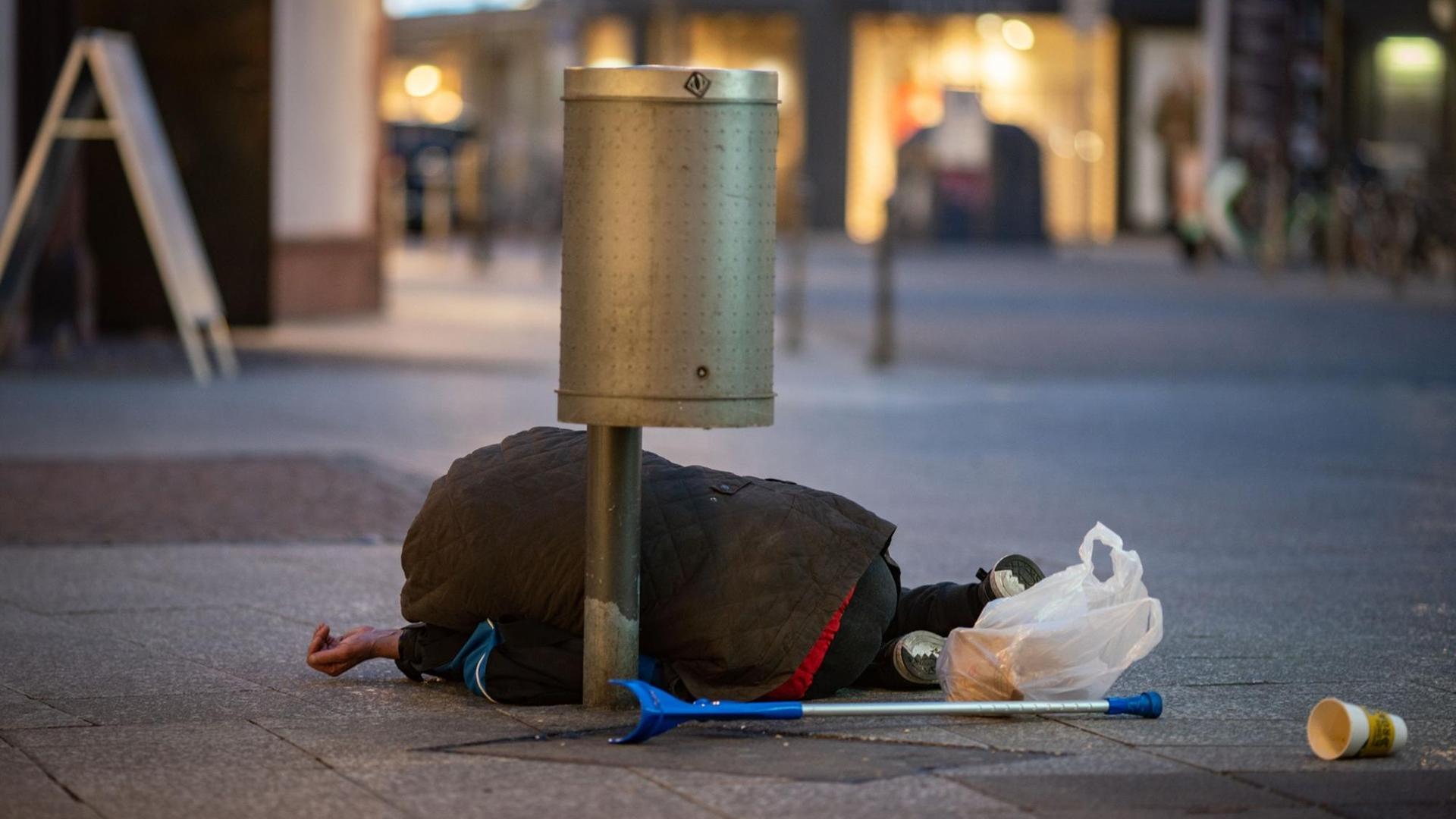 Ein Mann liegt in der Innenstadt unter einem Papierkorb, eine Hand zum Betteln geöffnet. Ein Mann liegt in der Innenstadt unter einem Papierkorb, eine Hand zum Betteln geöffnet.