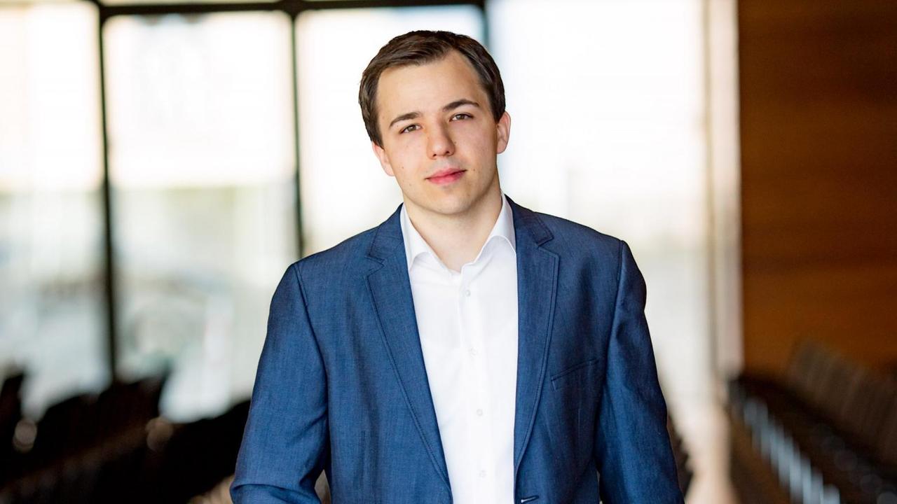 Ein Mann in weißem Hemd und blauem Sakko schaut frontal in die Kamera. Er hält den Kopf leicht nach links geneigt. Der Hintergrund ist unscharf abgebildet mit vielen Weiß- und einigen Brauntönen. Ein Mann in weißem Hemd und blauem Sakko schaut frontal in die Kamera. Er hält den Kopf leicht nach links geneigt. Der Hintergrund ist unscharf abgebildet mit vielen Weiß- und einigen Brauntönen.