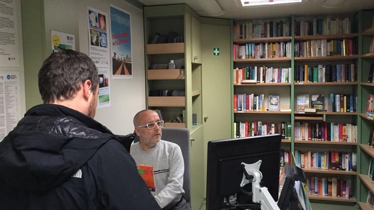 Mann am Computer, hinter ihm Bücherregale, vor ihm ein Mann der sich ein Buch ausleiht. Mann am Computer, hinter ihm Bücherregale, vor ihm ein Mann der sich ein Buch ausleiht.