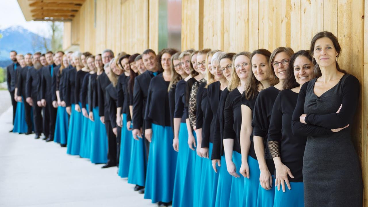 Der Chor steht in einer langen Schlange an einer Holzhausfront aufgestellt. Die Damen tragen dabei hellblaue Röcke und Oberteile aus Spitze. Der Chor steht in einer langen Schlange an einer Holzhausfront aufgestellt. Die Damen tragen dabei hellblaue Röcke und Oberteile aus Spitze.