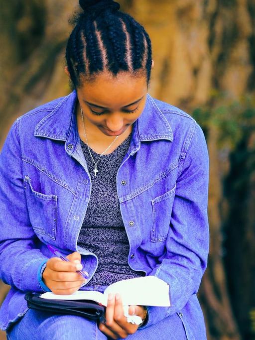 Eine Schwarze Frau sitzt hält einen Stift in der Hand und liest dabei in einem Buch.