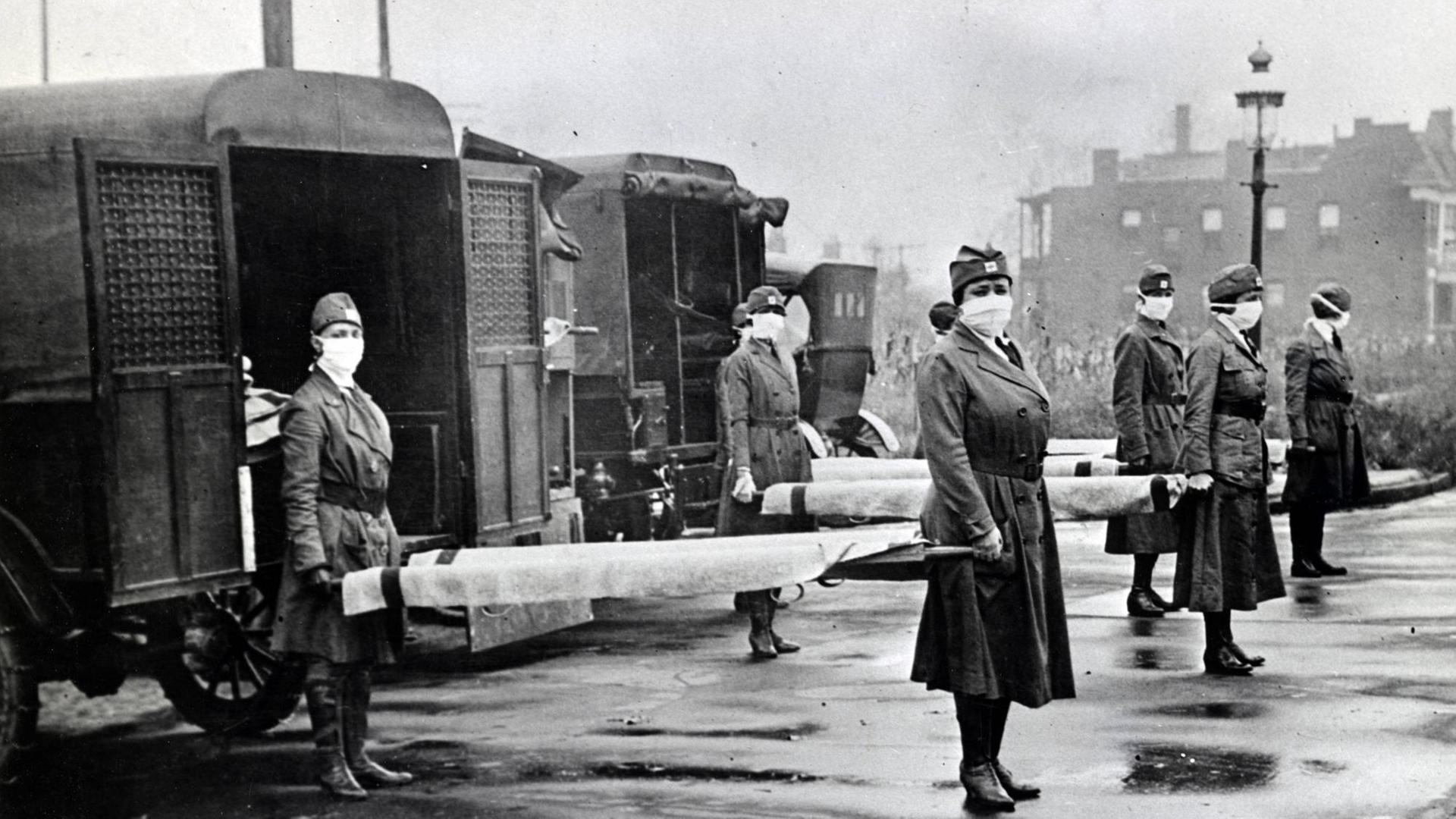 Schwarzweiß-Aufnahme um 1918: Frauen mit Mundschutz stehen mit Tragen neben Krankenwagen des Roten Kreuz in St. Louis. Schwarzweiß-Aufnahme um 1918: Frauen mit Mundschutz stehen mit Tragen neben Krankenwagen des Roten Kreuz in St. Louis.