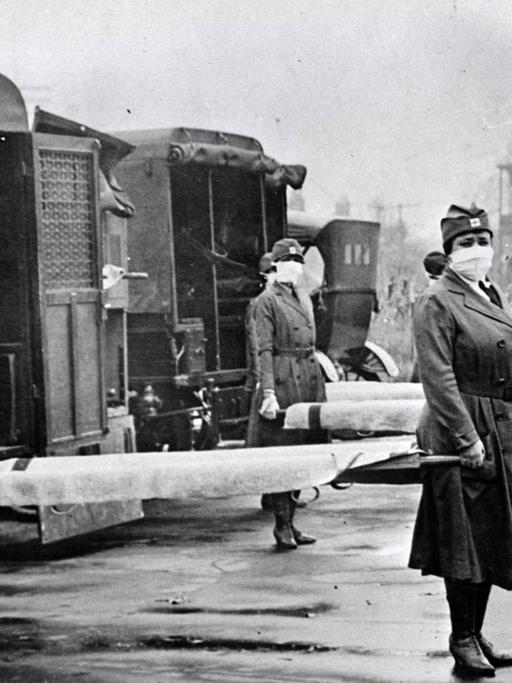 Schwarzweiß-Aufnahme um 1918: Frauen mit Mundschutz stehen mit Tragen neben Krankenwagen des Roten Kreuz in St. Louis.