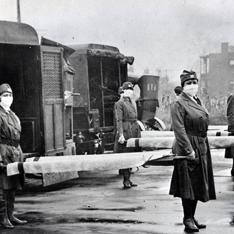 Schwarzweiß-Aufnahme um 1918: Frauen mit Mundschutz stehen mit Tragen neben Krankenwagen des Roten Kreuz in St. Louis.