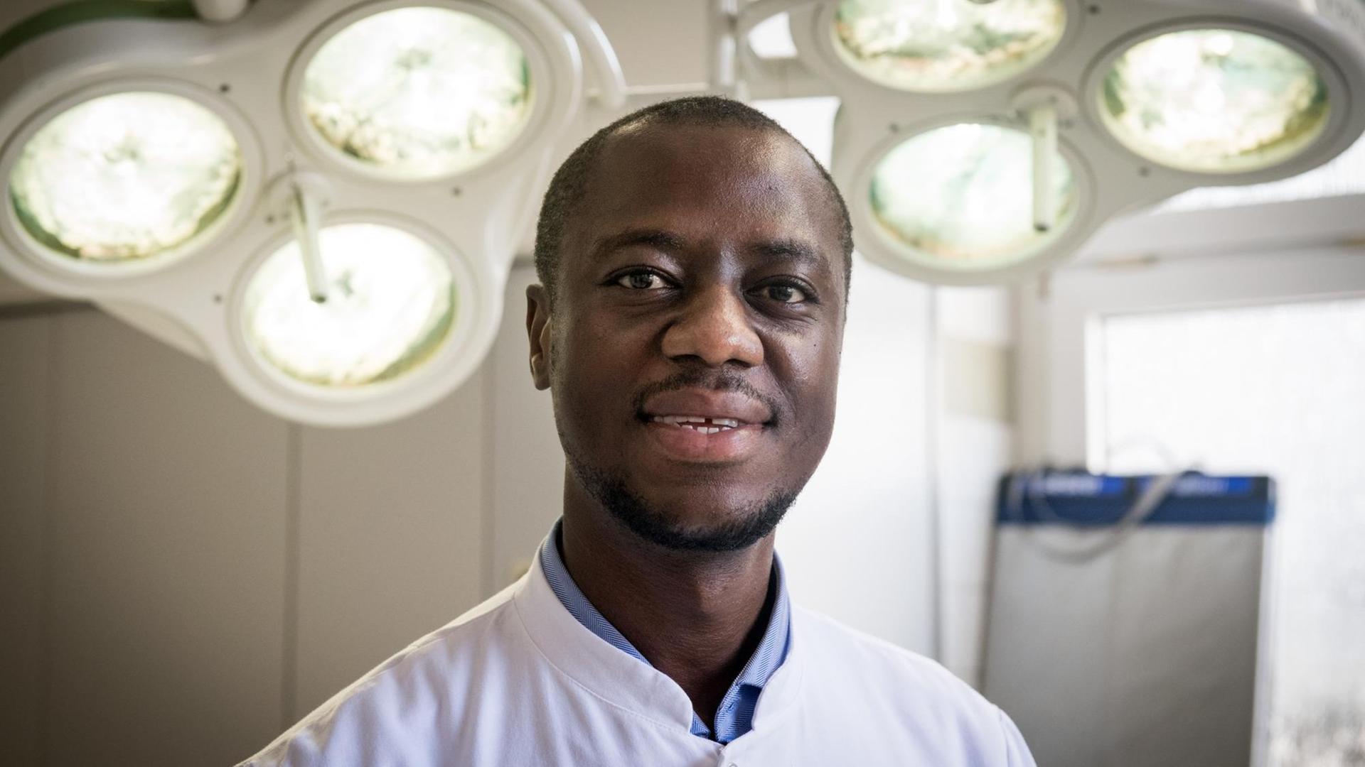 Der Arzt Samuel Samuel Okae trägt einen weißen Arztkittel. Er steht in einem Op-Saal und lächelt in die Kamera. Der Arzt Samuel Samuel Okae trägt einen weißen Arztkittel. Er steht in einem Op-Saal und lächelt in die Kamera.