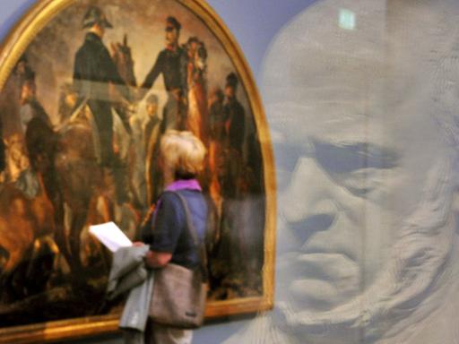 Eine Besucherin schaut sich das Gemälde "Blüchers Begegnung mit Wellington nach der Schlacht bei Belle-Alliance, 1858" von Adolph Menzel an. Im Vordergrund spiegelt sich die Gipsbüste Menzels, geschaffen von Reinhold Begas, in einer Glasscheibe.
