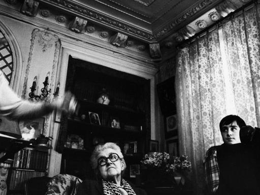 Die Schwarzweißfotografie zeigt einen herrschaftlich eingerichteten Wohnraum mit üppigem Stuck, geblümten Vorhängen, Kamin und Sesseln. Eine ältere Dame mit weißem Haar und dunkler Brille hört zu, während zwei Männer vermutlich musizieren.