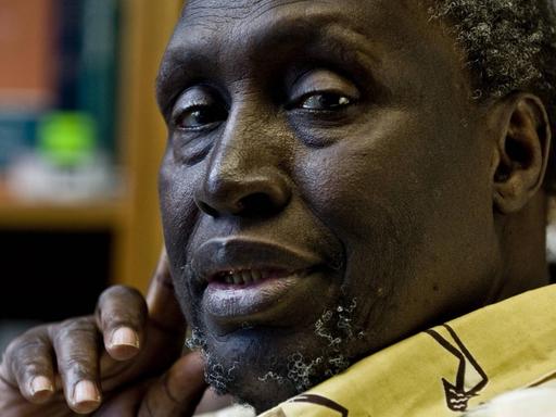 Portrait des kenianischen Schriftstellers Ngugi Wa Thiong o, der nach linke in die Kamera guckt und die rechte Hand mit Daumen und Zeigefinger an die Wange hält.
