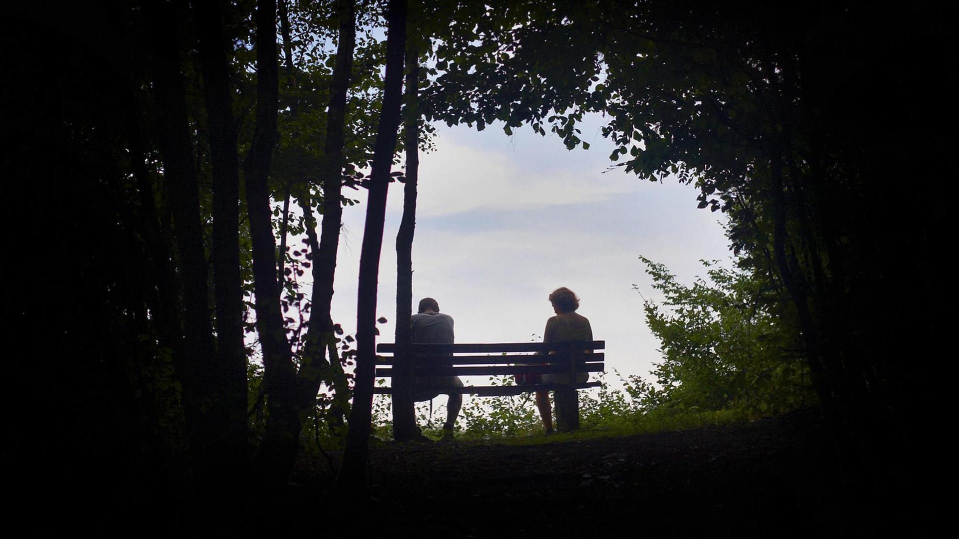 Ein Mann und eine Frau sitzen auf einer Bank zwischen Bäumen. Sie wirken erschöpft.