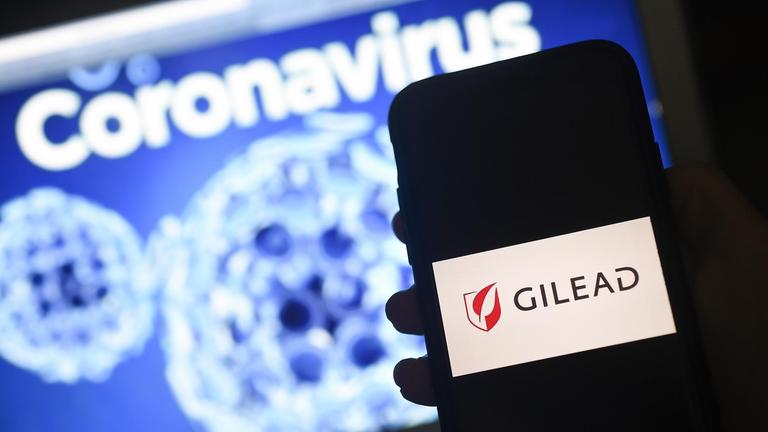 Das Gilead-Logo wird auf einem Smartphone neben einem Bildschirm mit einer Coronavirus-Grafik