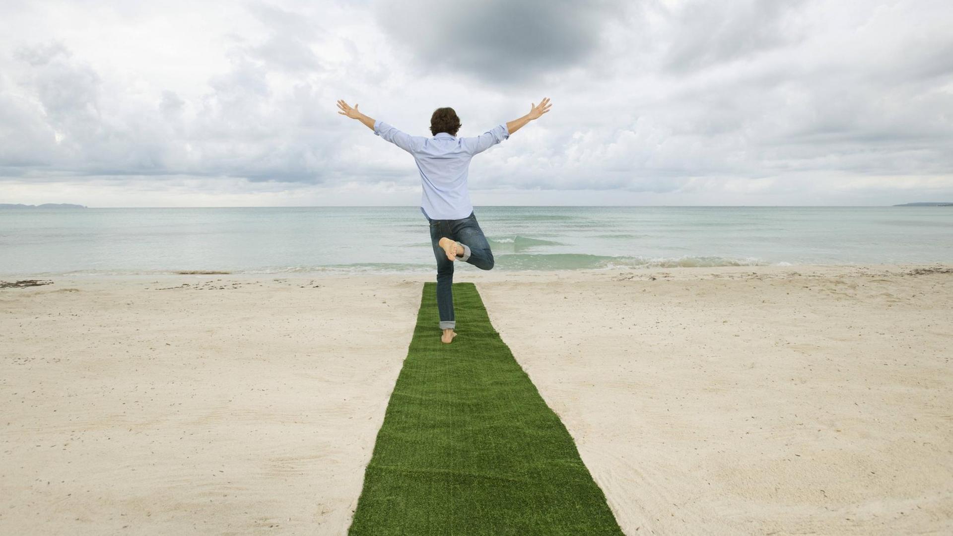 Ein Mann steht auf einem Bein auf einem grünen Teppich am Strand Ein Mann steht auf einem Bein auf einem grünen Teppich am Strand