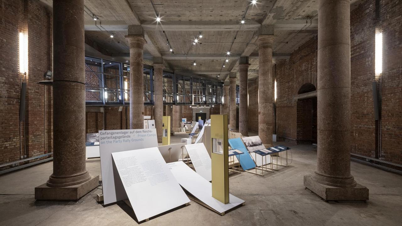 In einer Halle mit großen Säulen sind große Papptafeln aufgestellt, die die Geschichte des Nürnberger Arbeitslagers dokumentieren. In einer Halle mit großen Säulen sind große Papptafeln aufgestellt, die die Geschichte des Nürnberger Arbeitslagers dokumentieren.