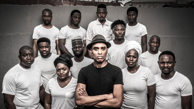 Elf afrikanische Männer und zwei Frauen stehen vor einer grauen Wand und blicken in die Kamera. Alle tragen weiße T-Shirts, nur der Mann in der Mitte trägt schwarz.