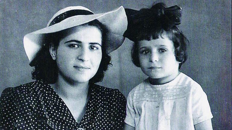 Eine Frau mit weißem Hut und großer Krempe sitzt neben ihrer sehr kleinen Tochter mit prächtiger Haarschleife.