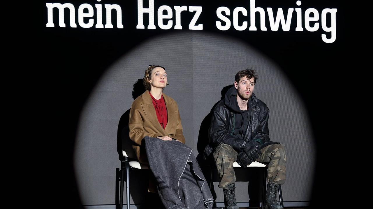 Foto von der Inszenierung: Ein Mann und eine Frau sitzen auf Stühlen auf der Bühne. Über ihren Köpfen ist der Schriftzug "Mein Herz schwieg" zu lesen. Foto von der Inszenierung: Ein Mann und eine Frau sitzen auf Stühlen auf der Bühne. Über ihren Köpfen ist der Schriftzug "Mein Herz schwieg" zu lesen.
